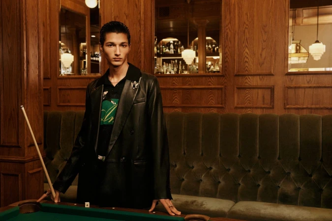A young man wearing a black leather jacket standing in front of a wood-paneled bar holding a billiard cue.