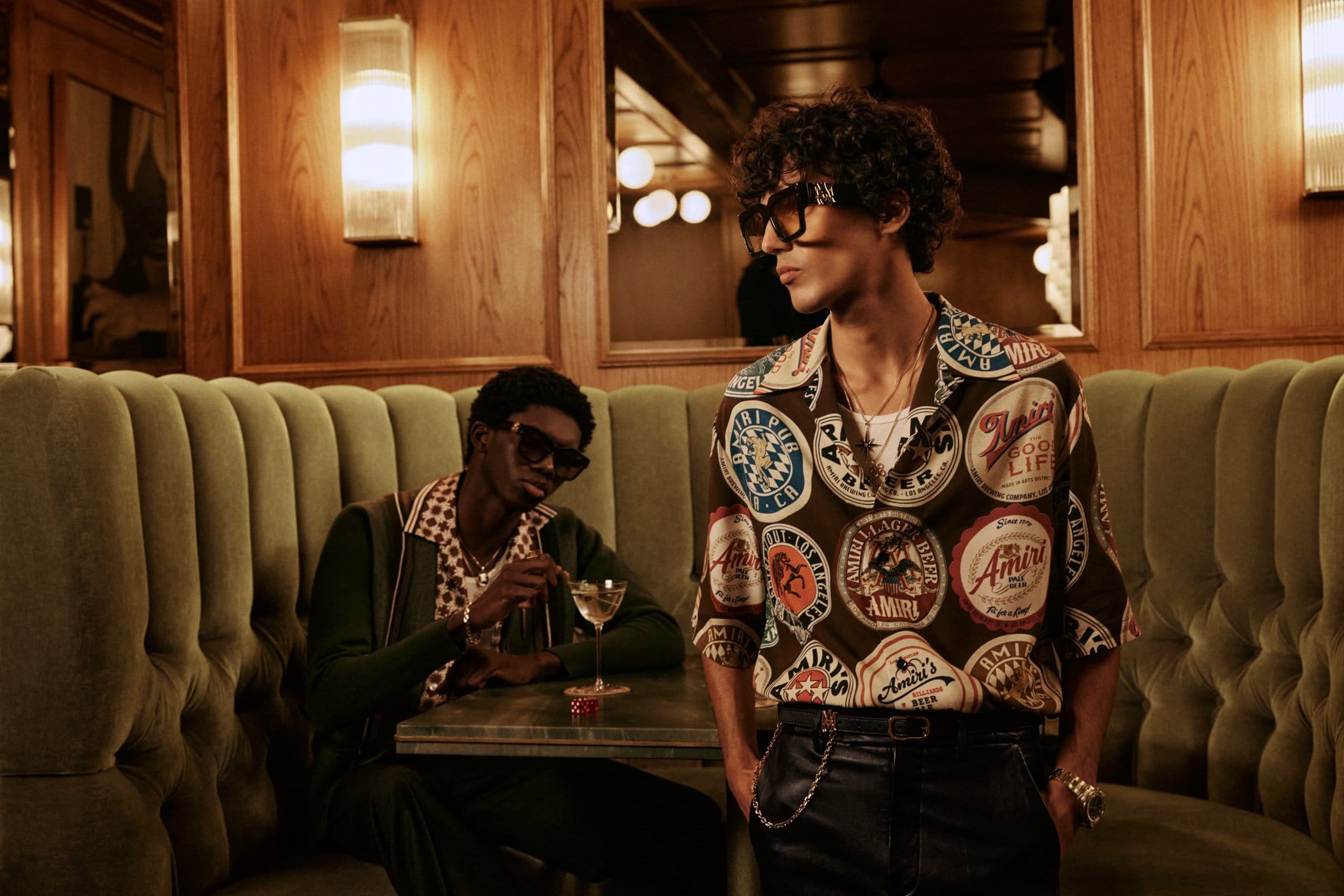 Two men seated in a plush green booth interior, one wearing a patterned button-down shirt and sunglasses