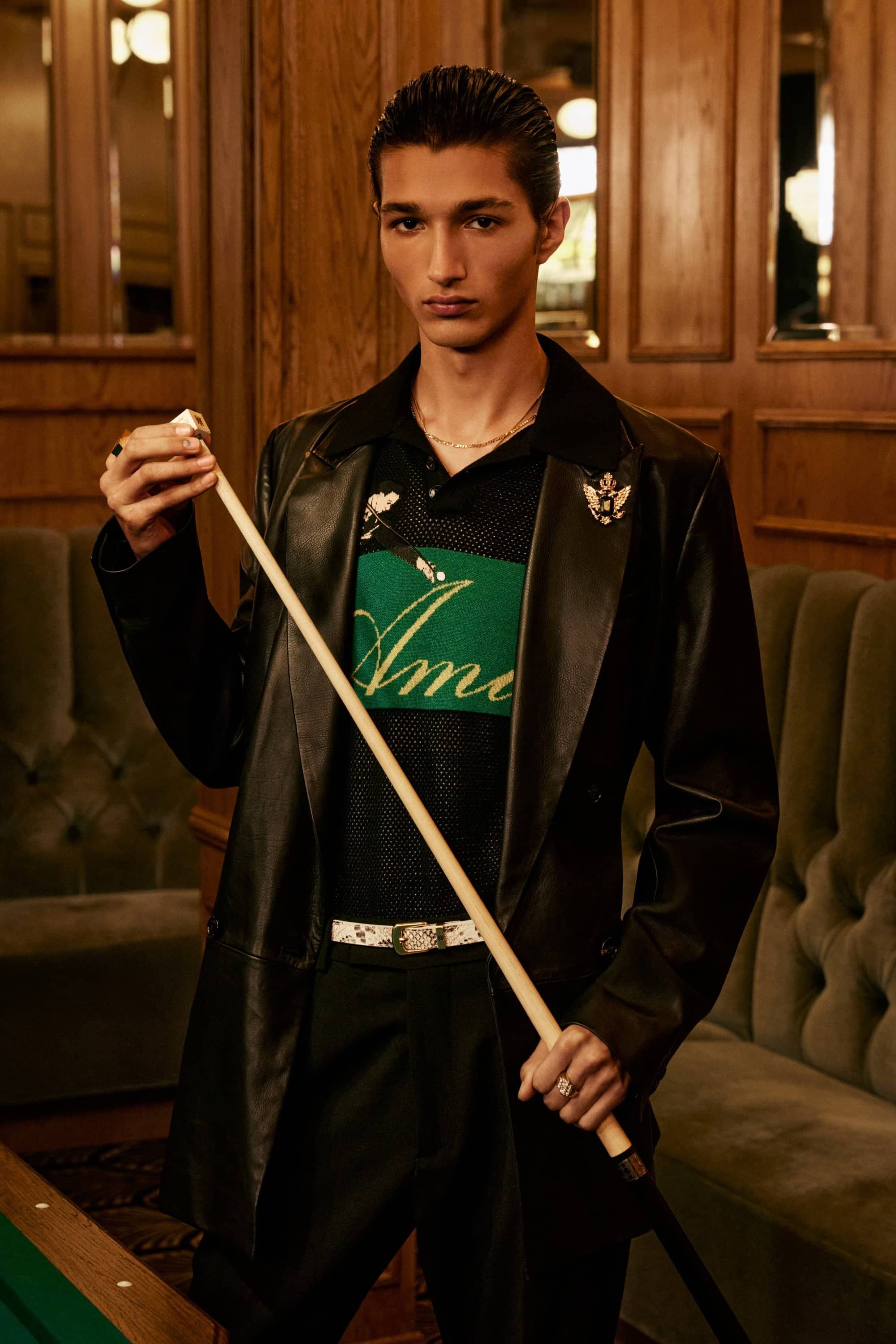 A young male model wearing a dark leather blazer and green polo shirt holds a billiard cue inside a wood-paneled lounge.
