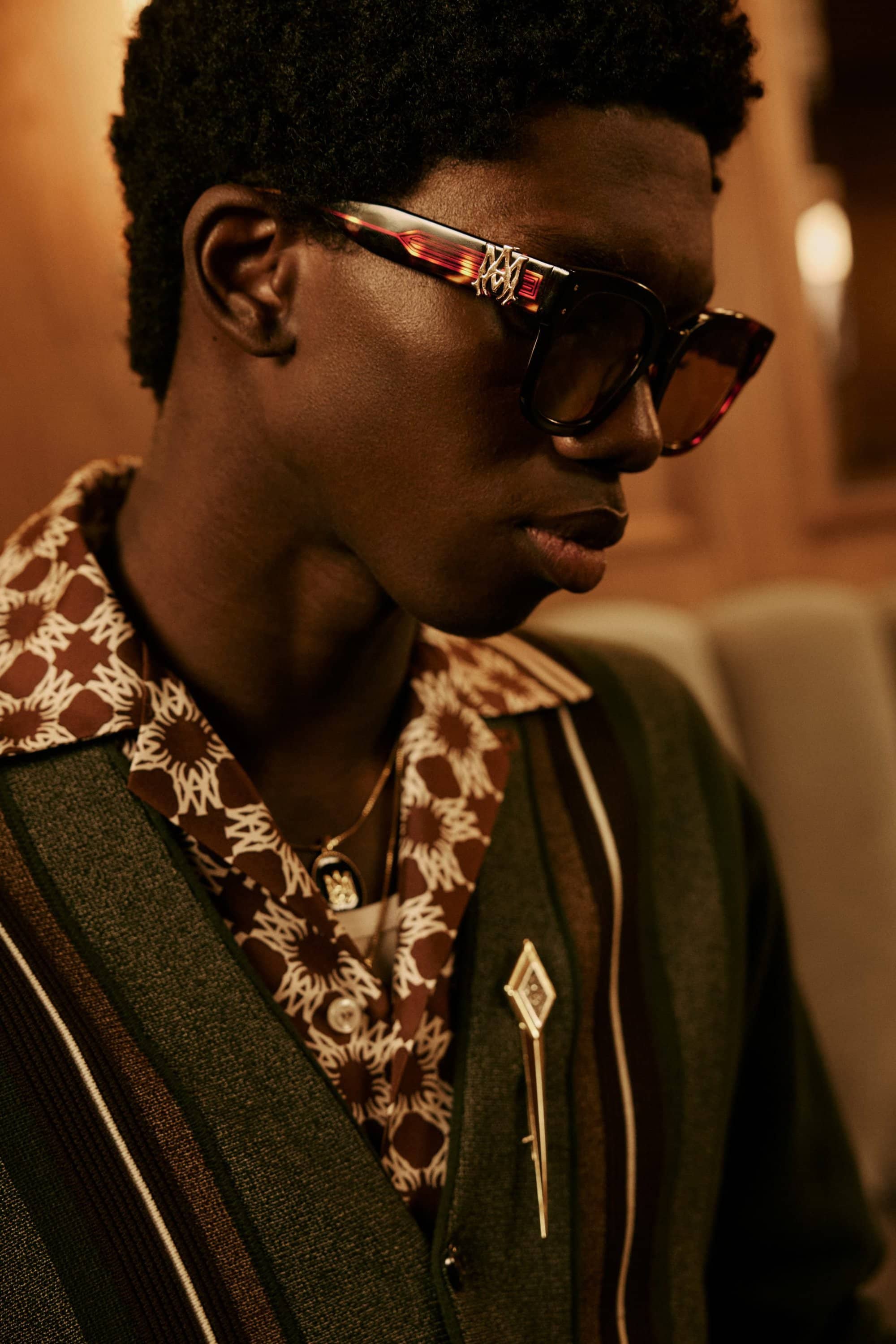 Portrait of a man wearing dark sunglasses, patterned shirt, and green cardigan with a brooch for Amiri Pre-Spring 2025 campaign.