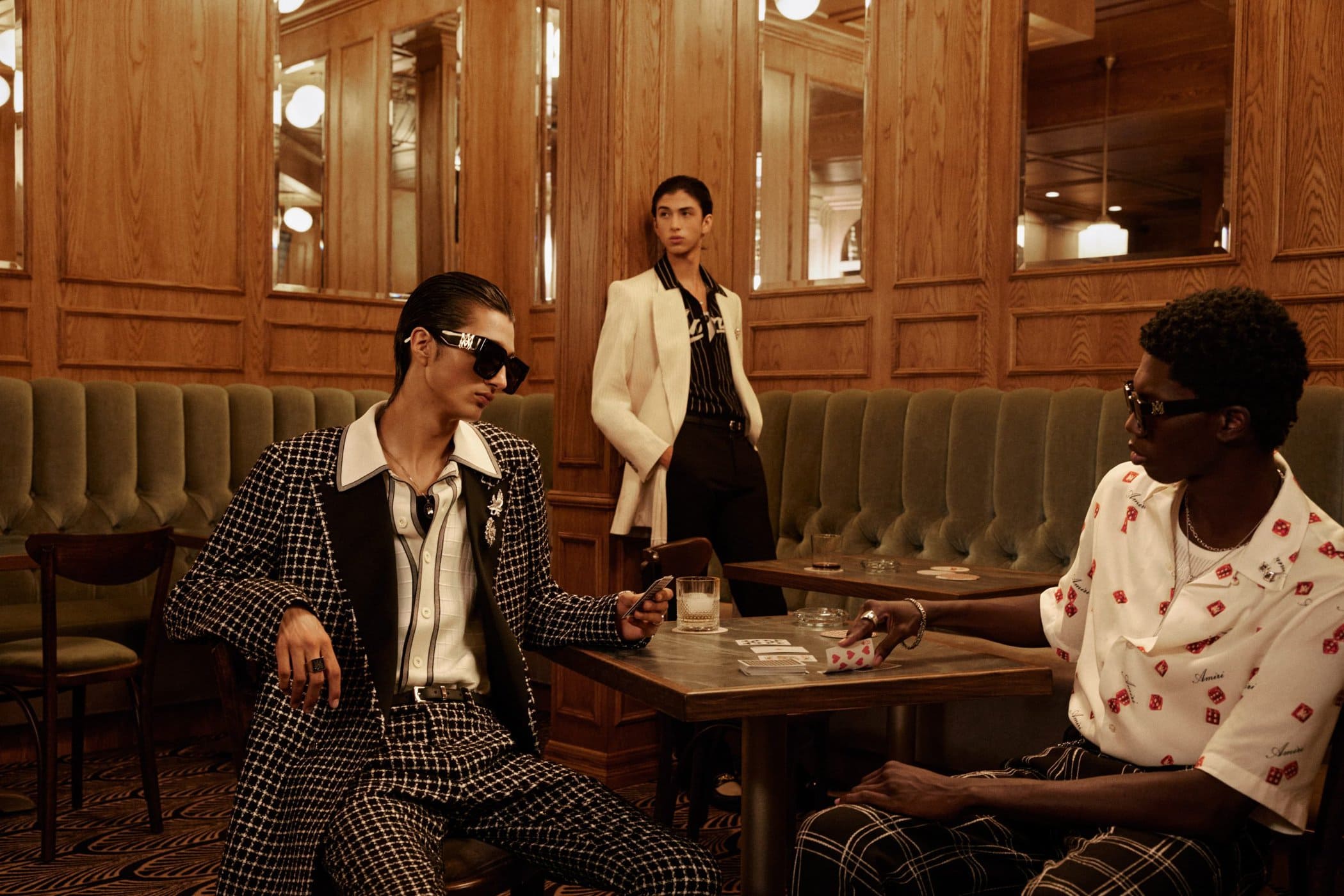 Three models wearing designer menswear inside a wood-paneled lounge with green leather booths for Amiri campaign.