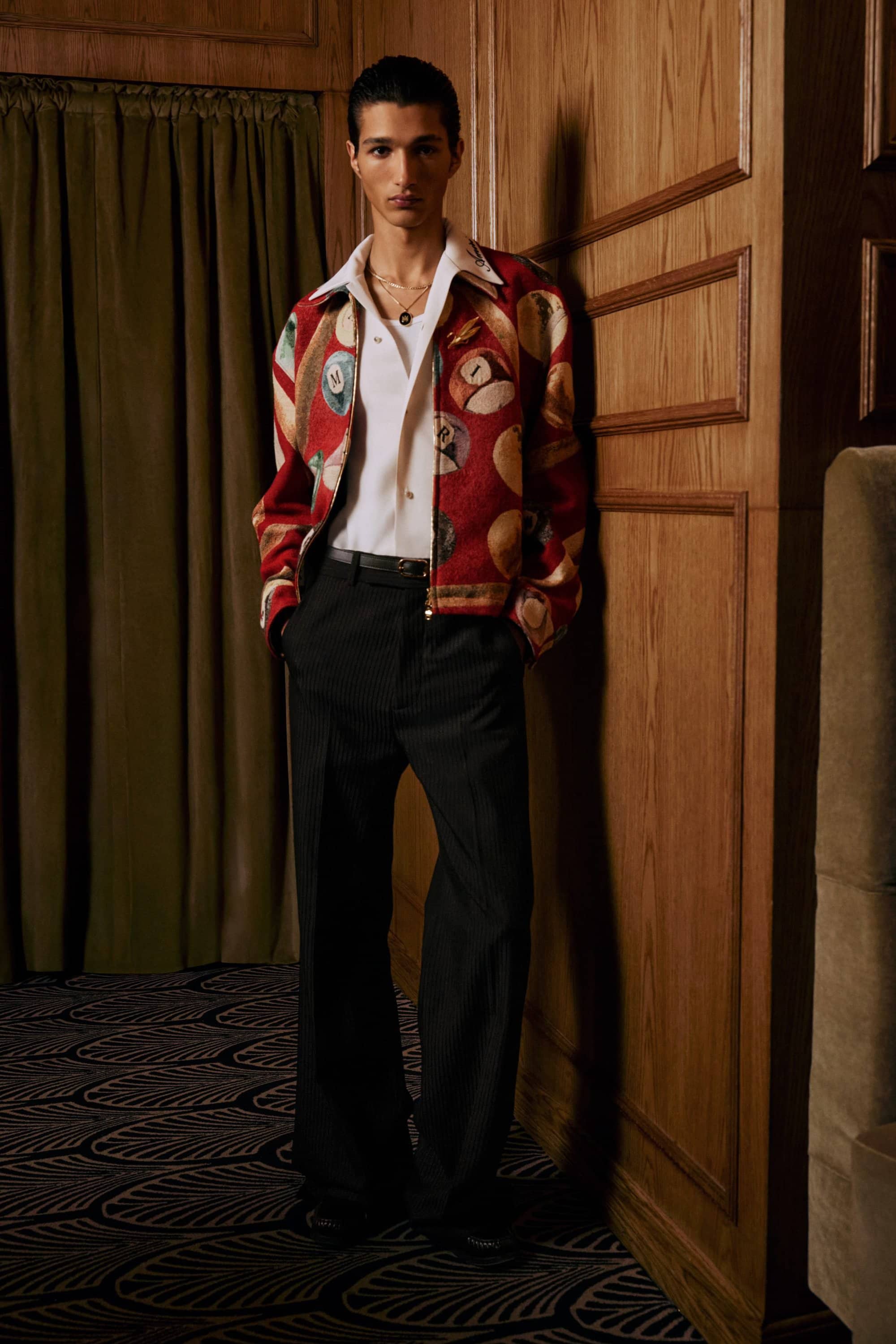 A man posing against wood paneling wearing a patterned red jacket, white shirt, and dark trousers.