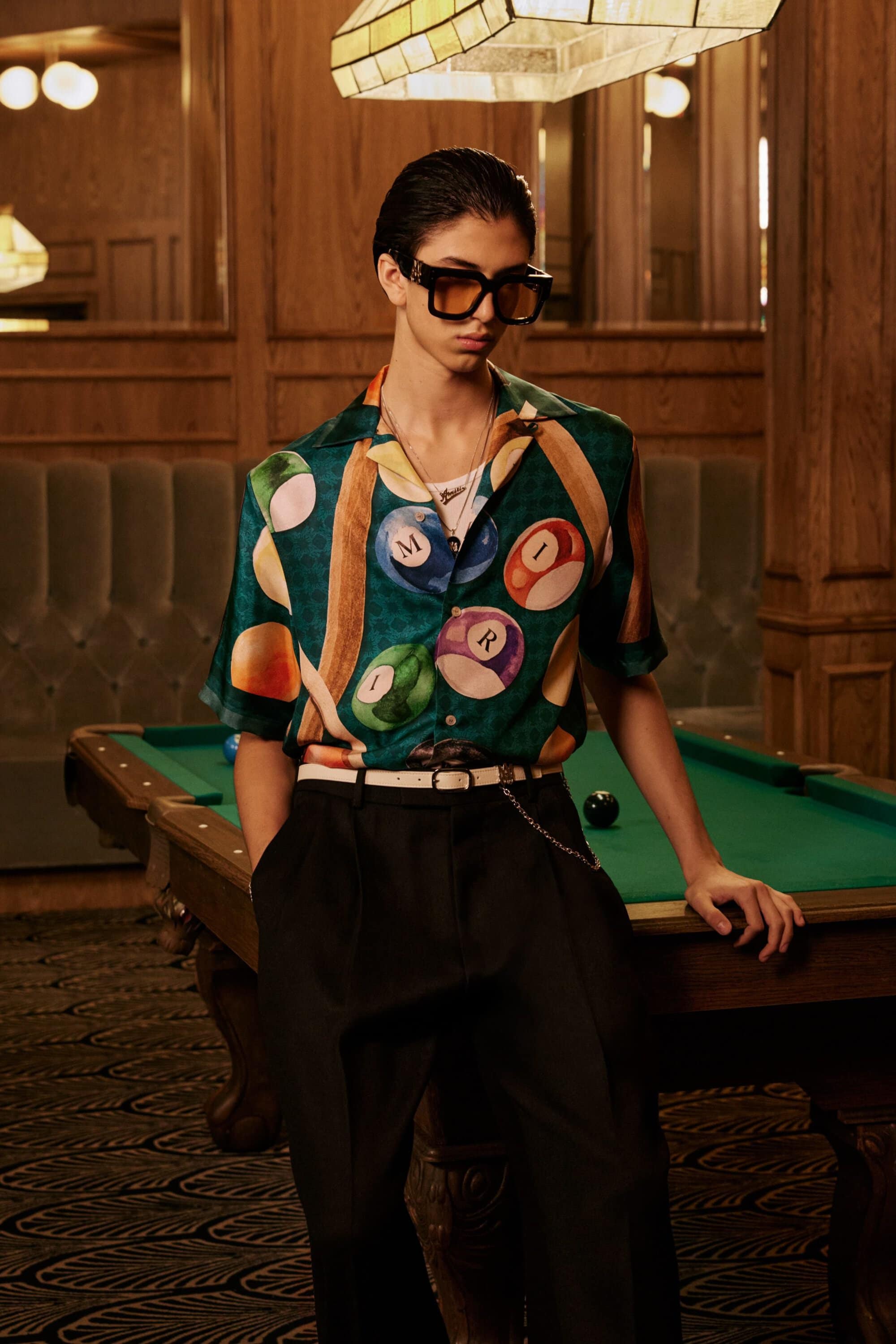 A man wearing oversized sunglasses and a patterned billiard ball shirt leans against a pool table in a wood-paneled room.