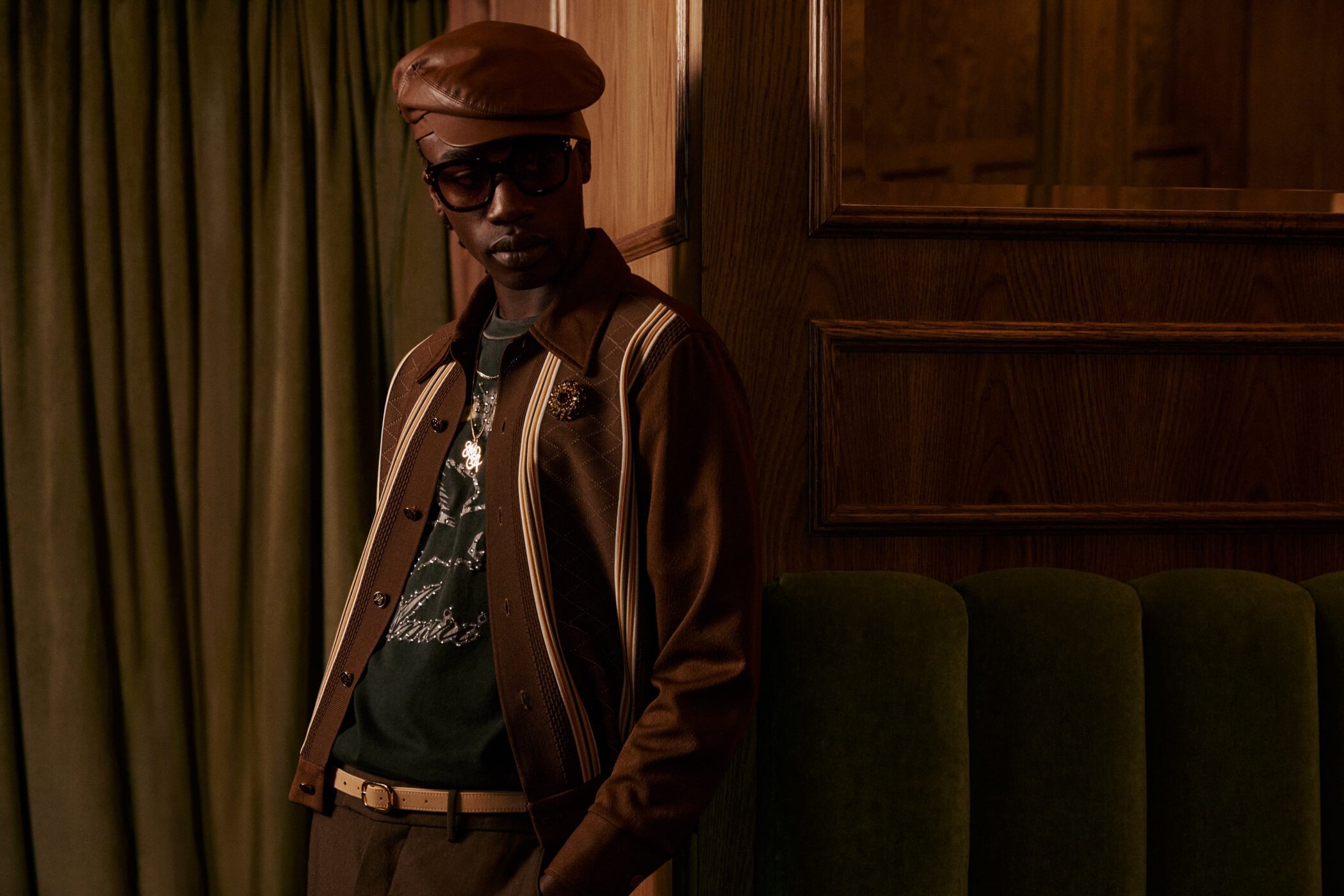 Man wearing a brown leather cap, sunglasses, and a brown jacket leaning against a dark wood wall with green velvet seating.