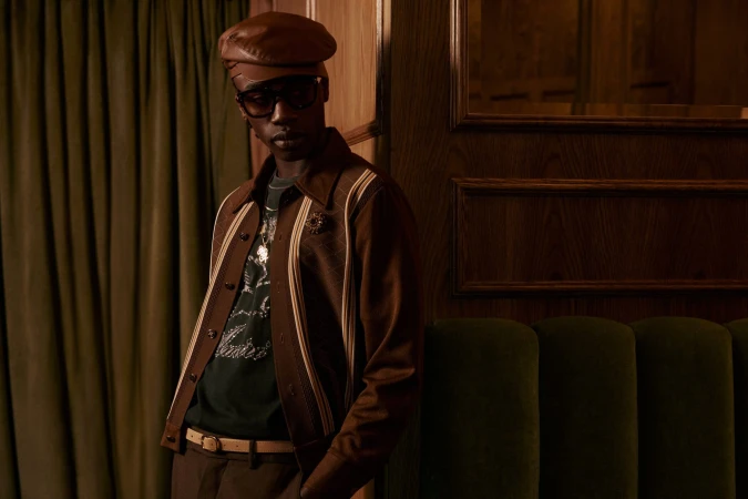 Man wearing a brown leather cap, sunglasses, and a brown jacket leaning against a dark wood wall with green velvet seating.