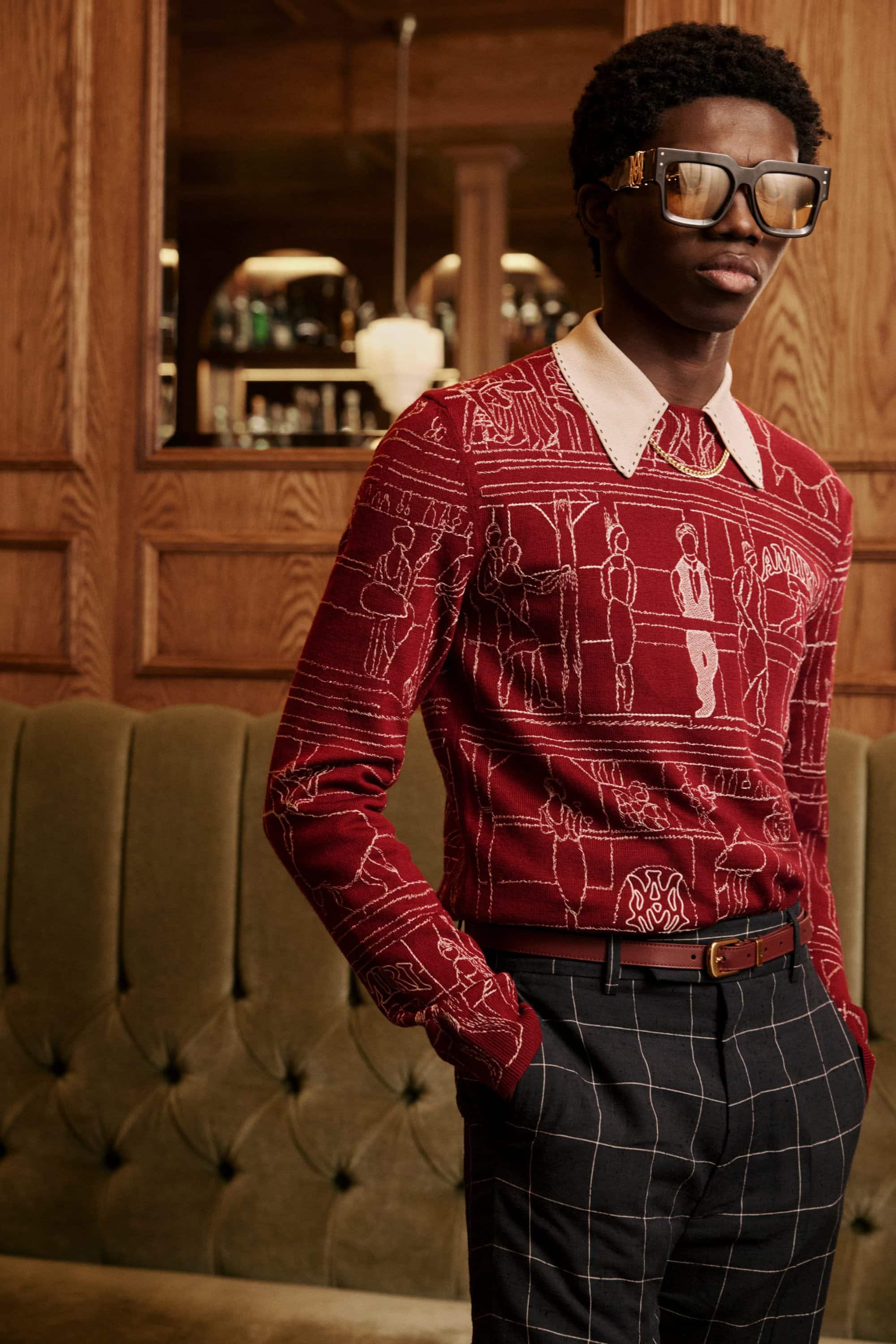 Man wearing graphic red patterned knit sweater and dark plaid trousers standing against a wood-paneled interior.