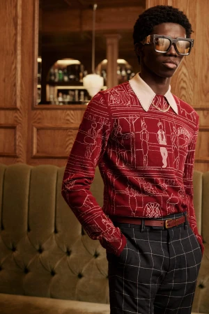 Man wearing graphic red patterned knit sweater and dark plaid trousers standing against a wood-paneled interior.