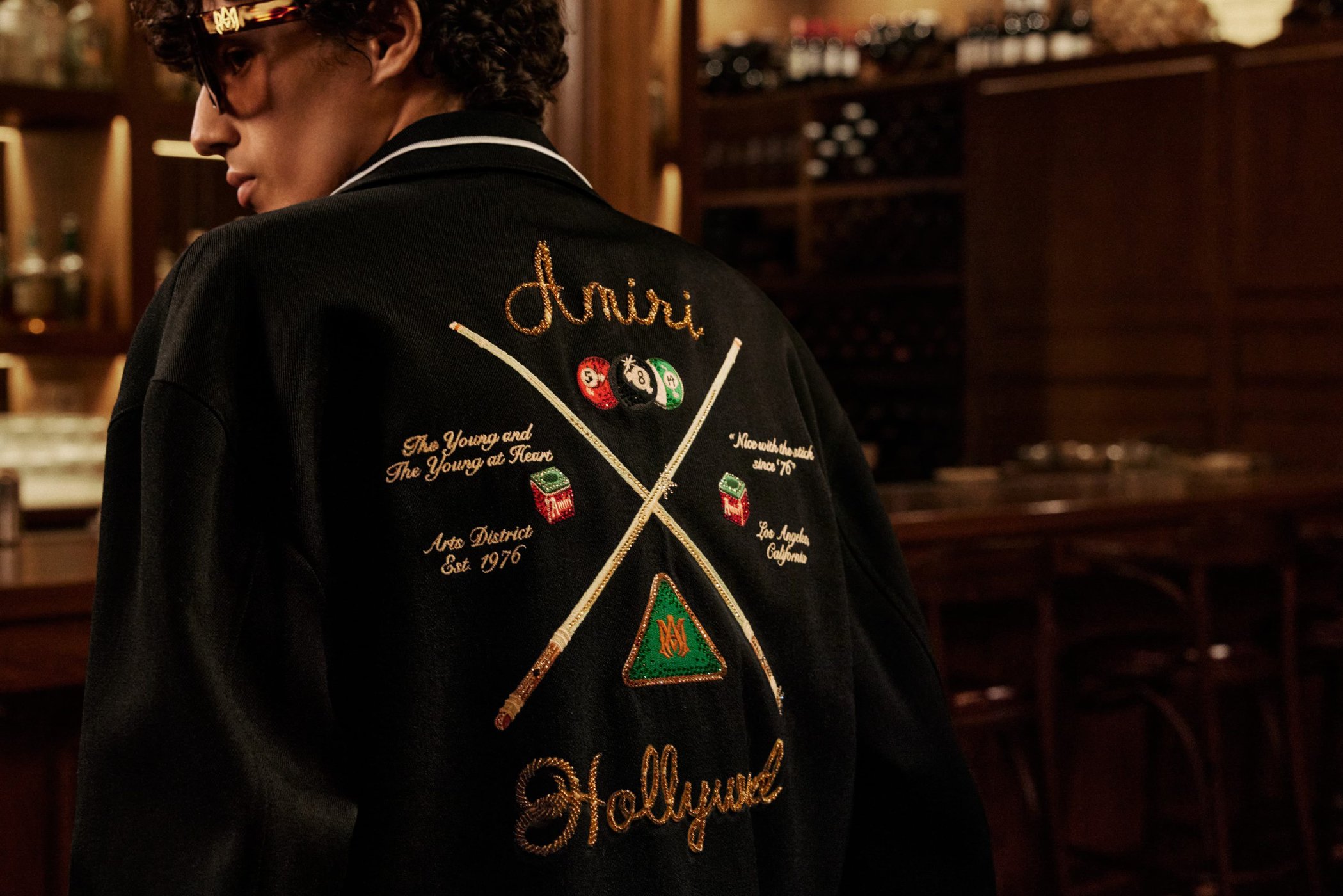 Model wearing a black jacket with detailed embroidery on the back in a dimly lit bar interior