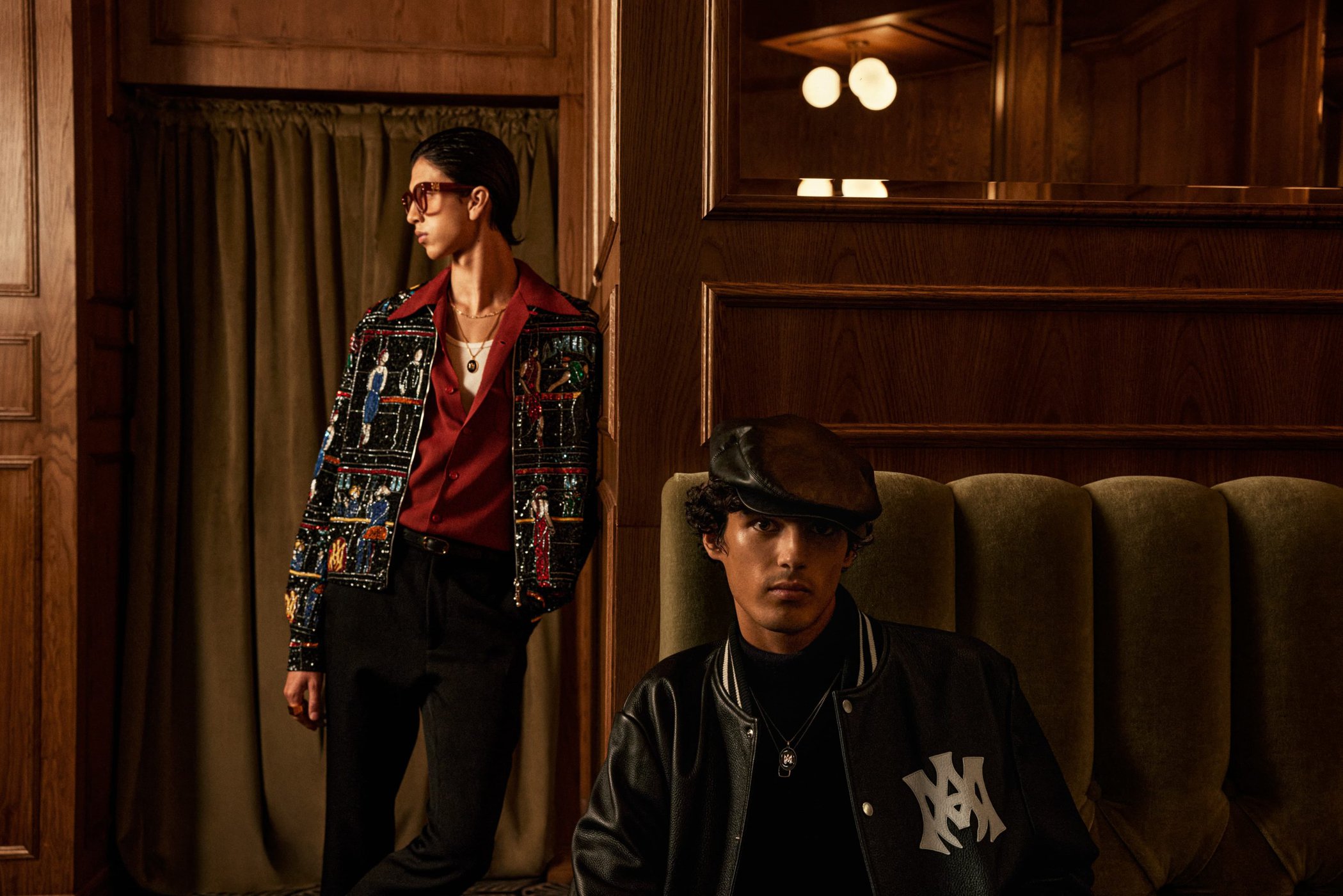 Two male models in an interior setting, one standing wearing an embellished jacket and red shirt, one seated on a green tufted sofa.
