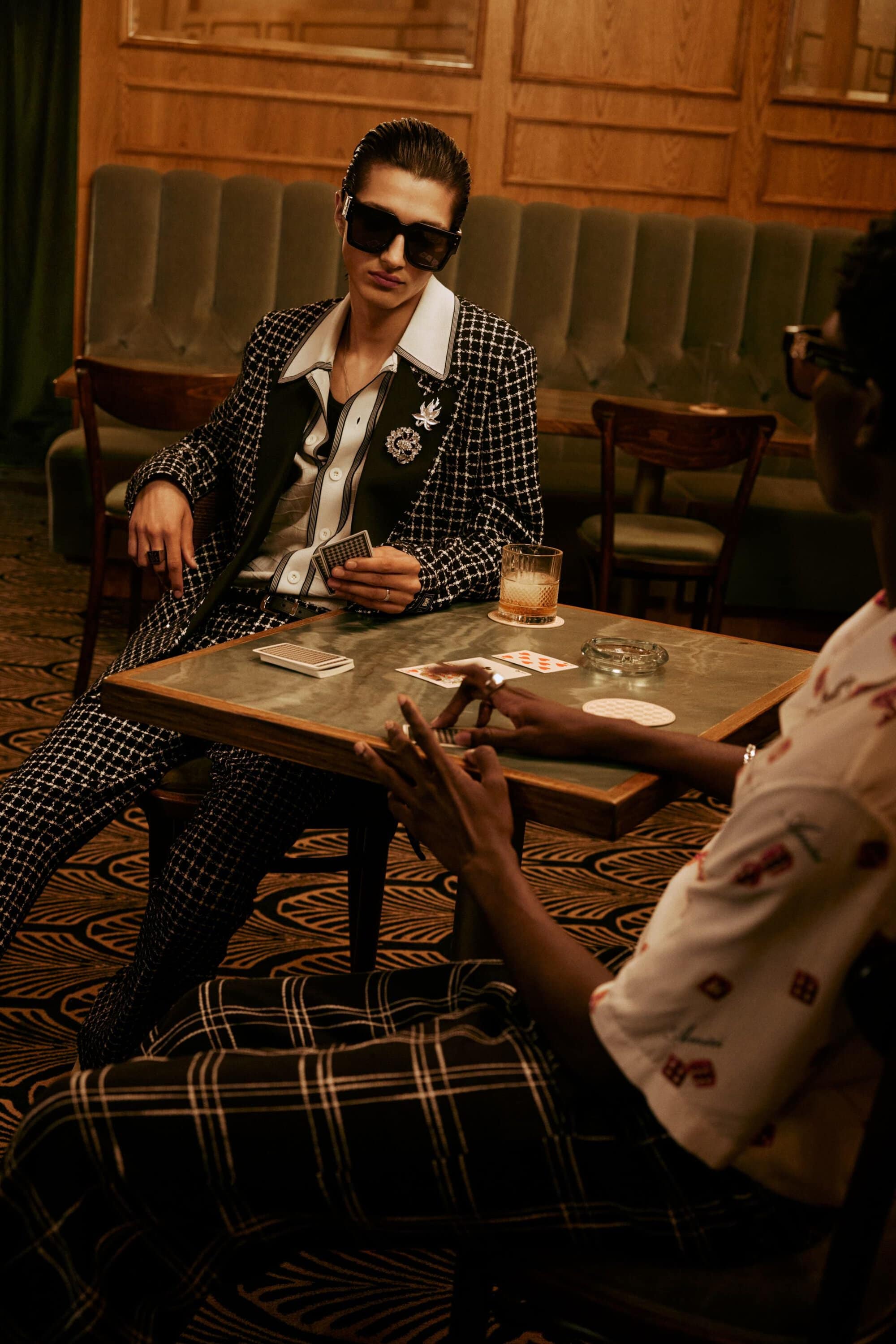 Two models seated at a wooden table playing cards in an intimate bar setting for the Amiri Pre-Spring 2025 campaign.