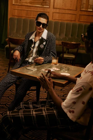 Two models seated at a wooden table playing cards in an intimate bar setting for the Amiri Pre-Spring 2025 campaign.