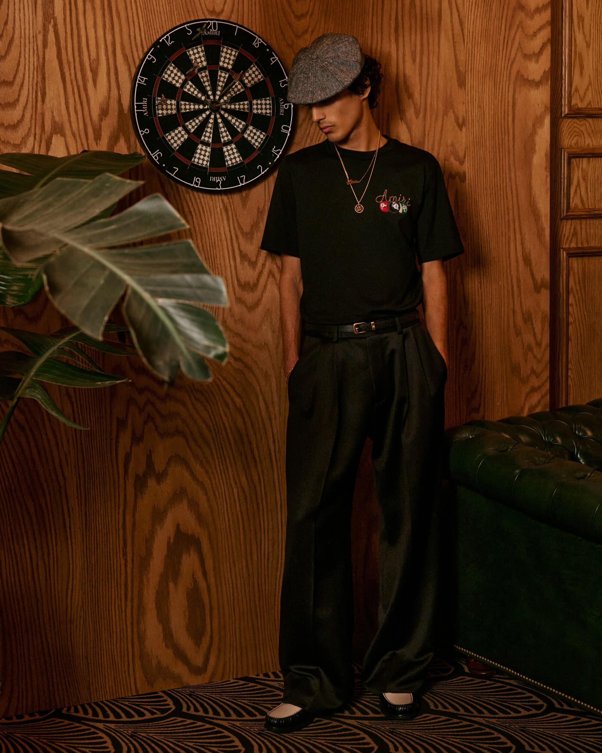 Man wearing a black t-shirt, dark trousers, and a flat cap standing against a wood-paneled wall next to a dartboard.