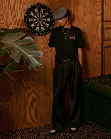 Man wearing a black t-shirt, dark trousers, and a flat cap standing against a wood-paneled wall next to a dartboard.