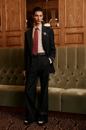 Man wearing a dark pinstripe suit and patterned red tie standing in an interior with wood paneling and a tufted green velvet banquette.