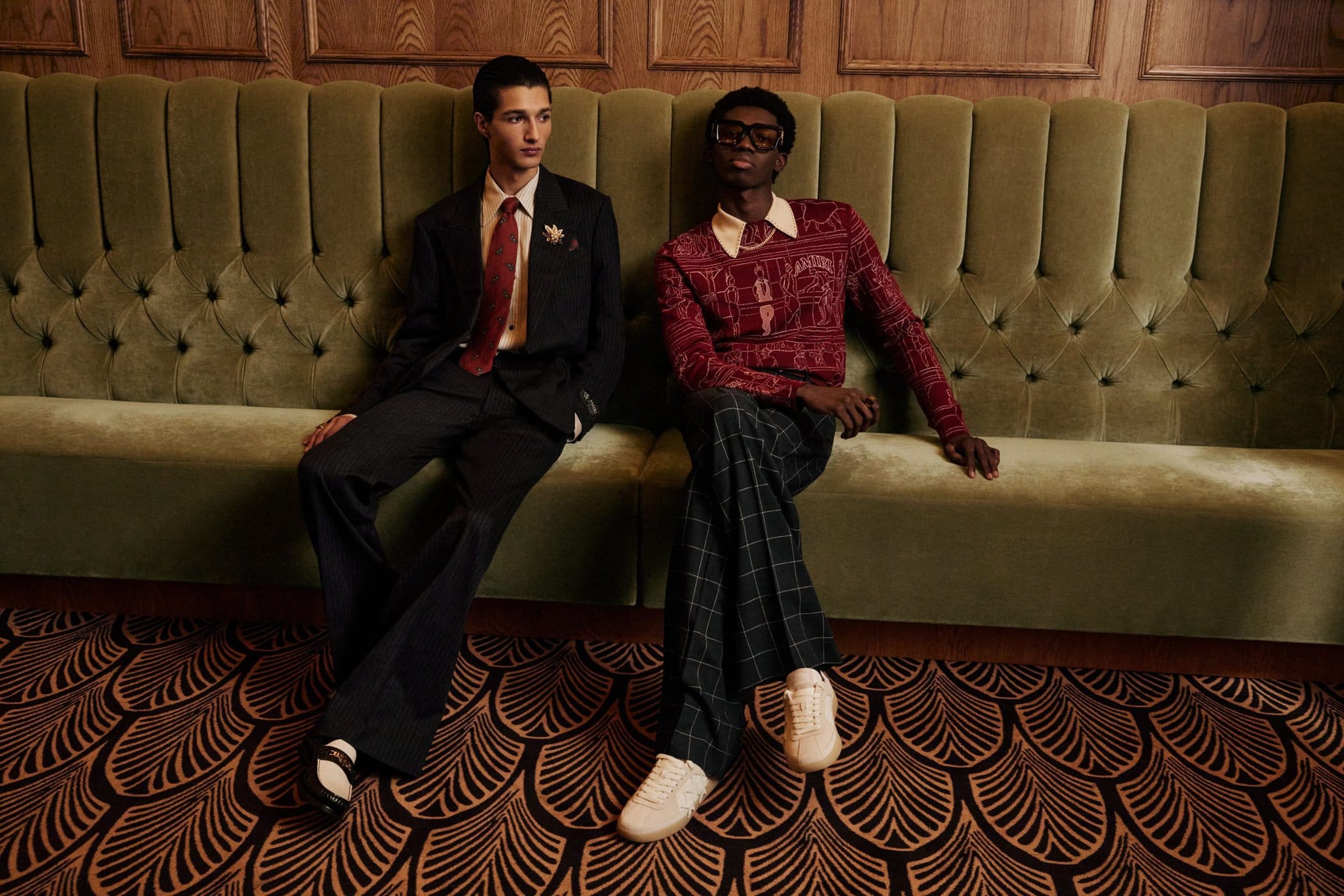 Two men seated together on a large green tufted velvet banquette in an interior setting for Amiri Pre-Spring 2025 campaign.