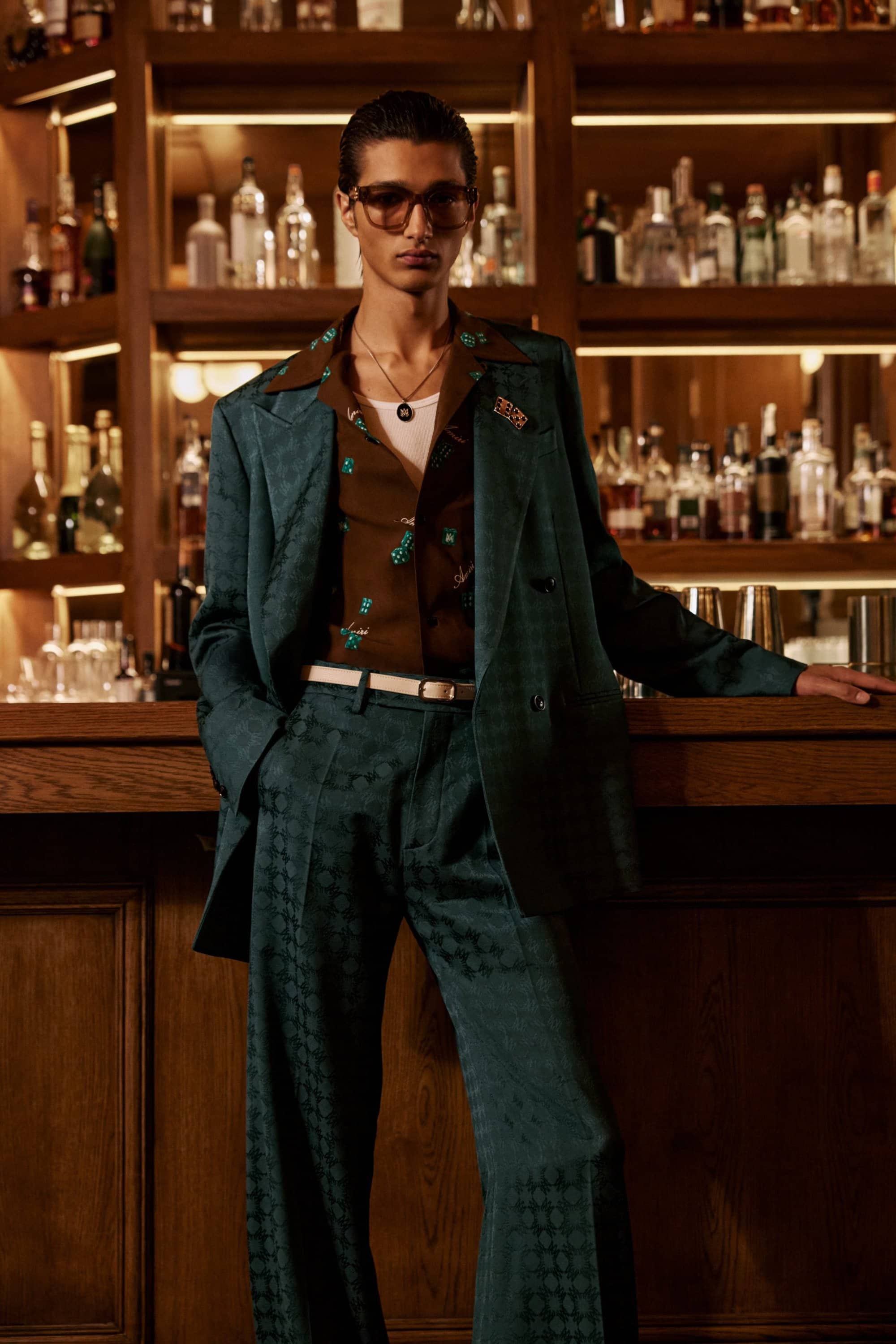 Model wearing a dark green suit and brown patterned shirt posing inside a bar for the Amiri Pre-Spring 2025 campaign.