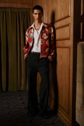 A man posing against wood paneling wearing a patterned red jacket, white shirt, and dark trousers.