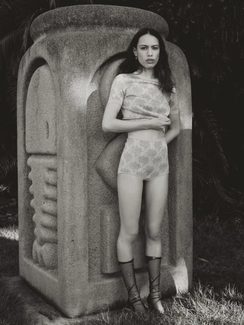 Young woman in patterned top and high-waisted shorts poses next to a carved stone sculpture in an outdoor setting with dark foliage.