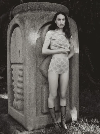 Young woman in patterned top and high-waisted shorts poses next to a carved stone sculpture in an outdoor setting with dark foliage.