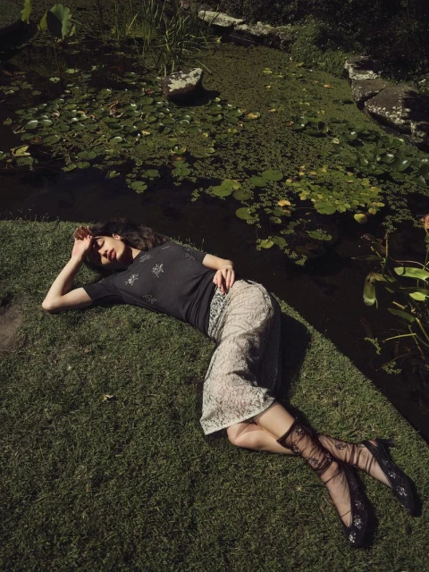 Woman rests on grass near a pond in a black top, floral skirt, and sheer patterned socks, bathed in dappled sunlight on a sunny day.