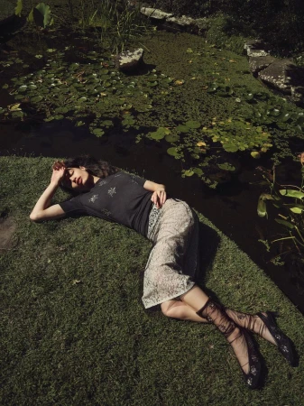 Woman rests on grass near a pond in a black top, floral skirt, and sheer patterned socks, bathed in dappled sunlight on a sunny day.