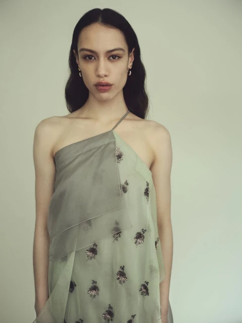 Woman wearing a draped, one-shoulder dress with a floral pattern, posing in a studio with soft, neutral lighting for a fashion editorial shot.