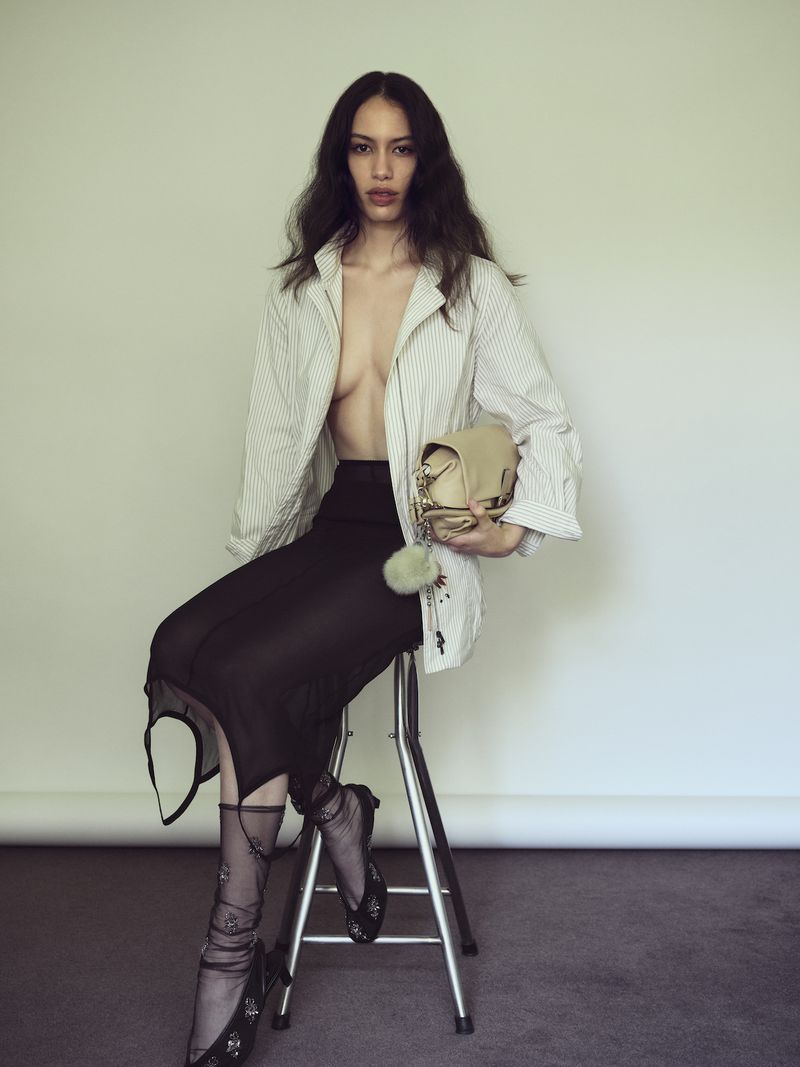Fashion portrait of an adult woman in a striped shirt and sheer black skirt sitting on a stool, holding a purse in a studio.