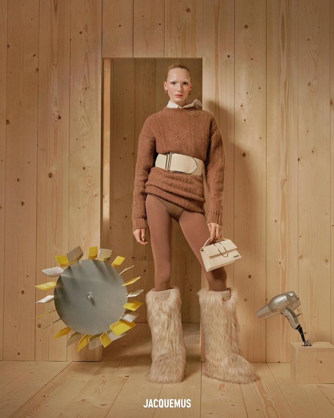 Model wearing a brown sweater, white belt, and furry boots holding a small white handbag against a wood panel wall