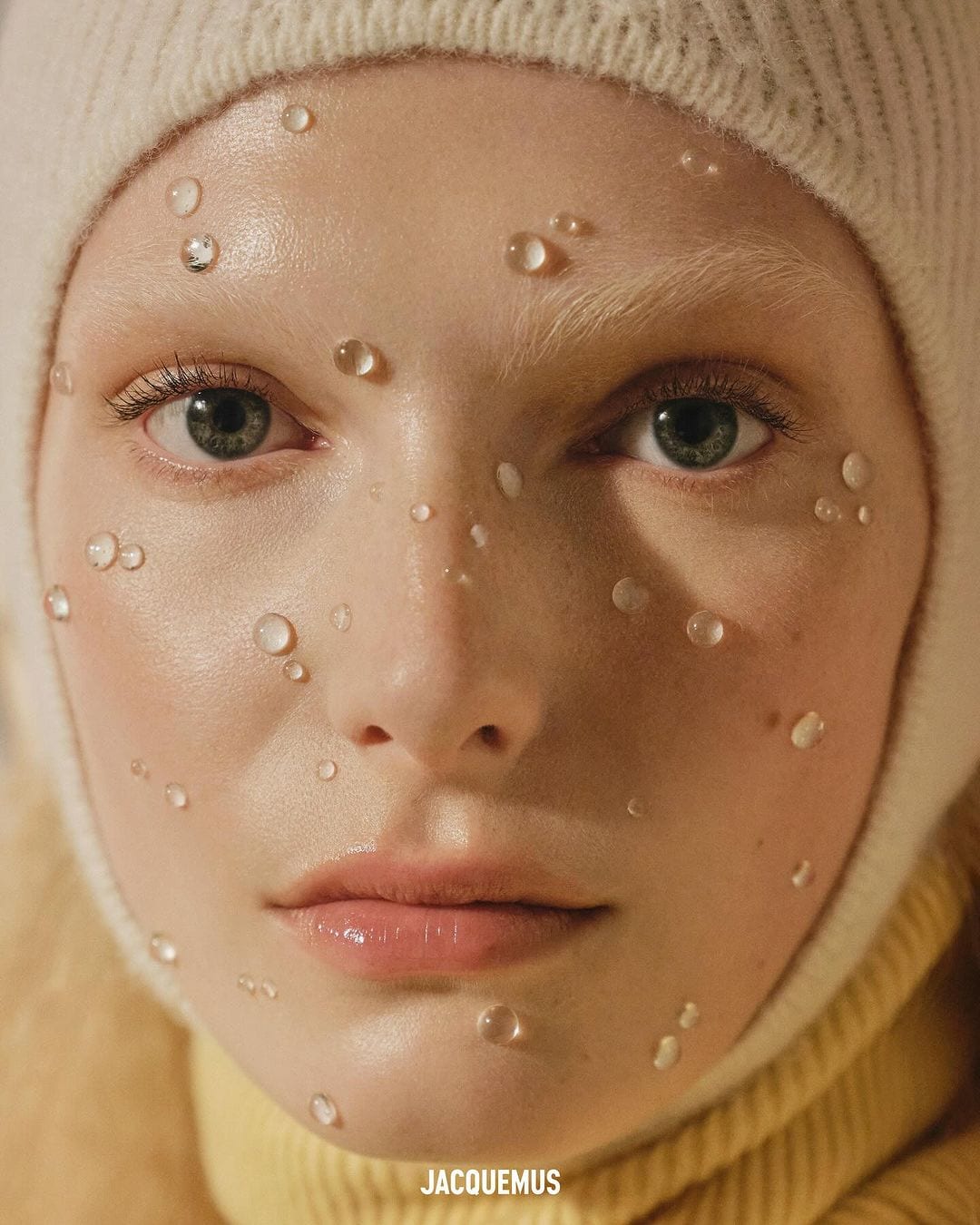 Close-up portrait of a model wearing a white knit cap with water droplets on her face for Jacquemus.