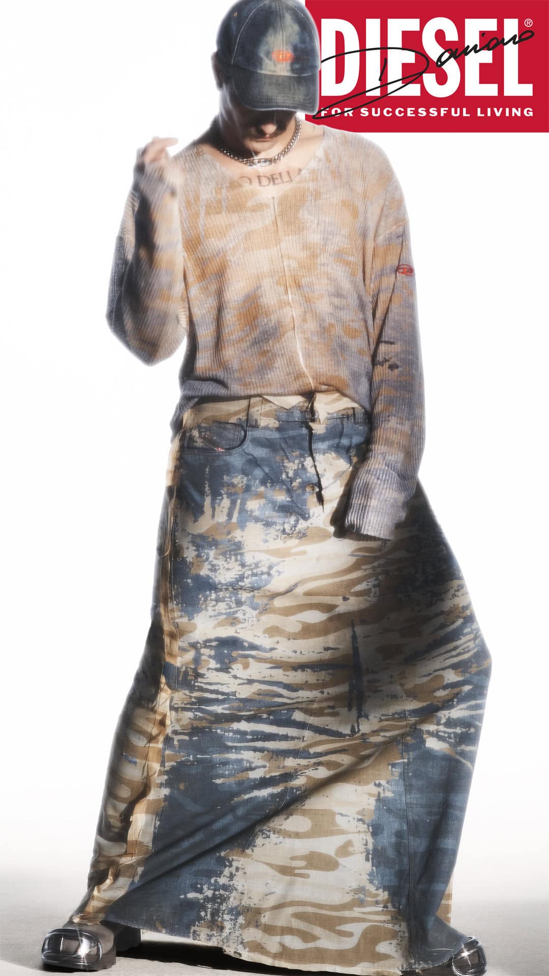 Damiano David in a long camo skirt and distressed knit sweater wearing a cap against a white background for Diesel Fall 2024