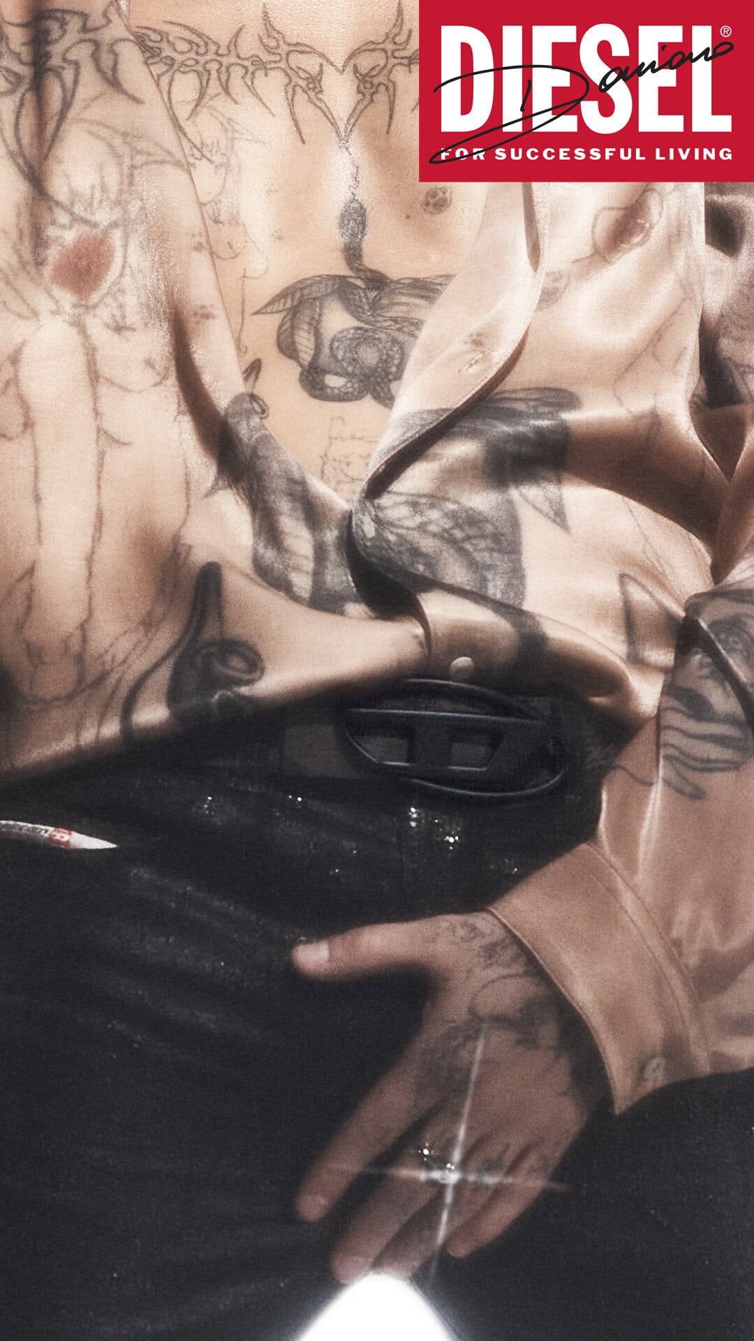 Close-up of Damiano David with torso tattoos wearing an open satin shirt and dark trousers for Diesel