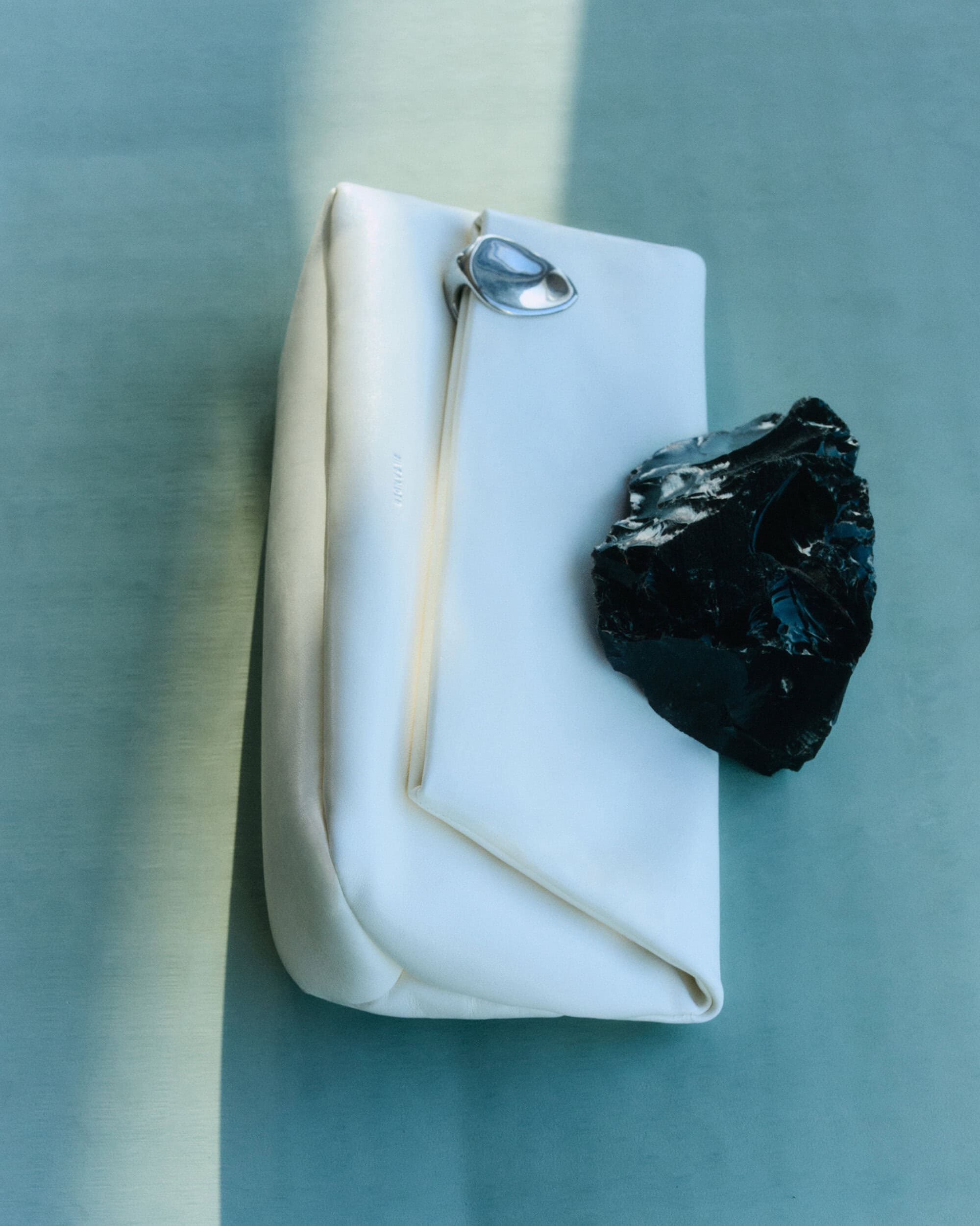 White leather clutch bag with silver clasp resting on a reflective surface alongside a dark rock