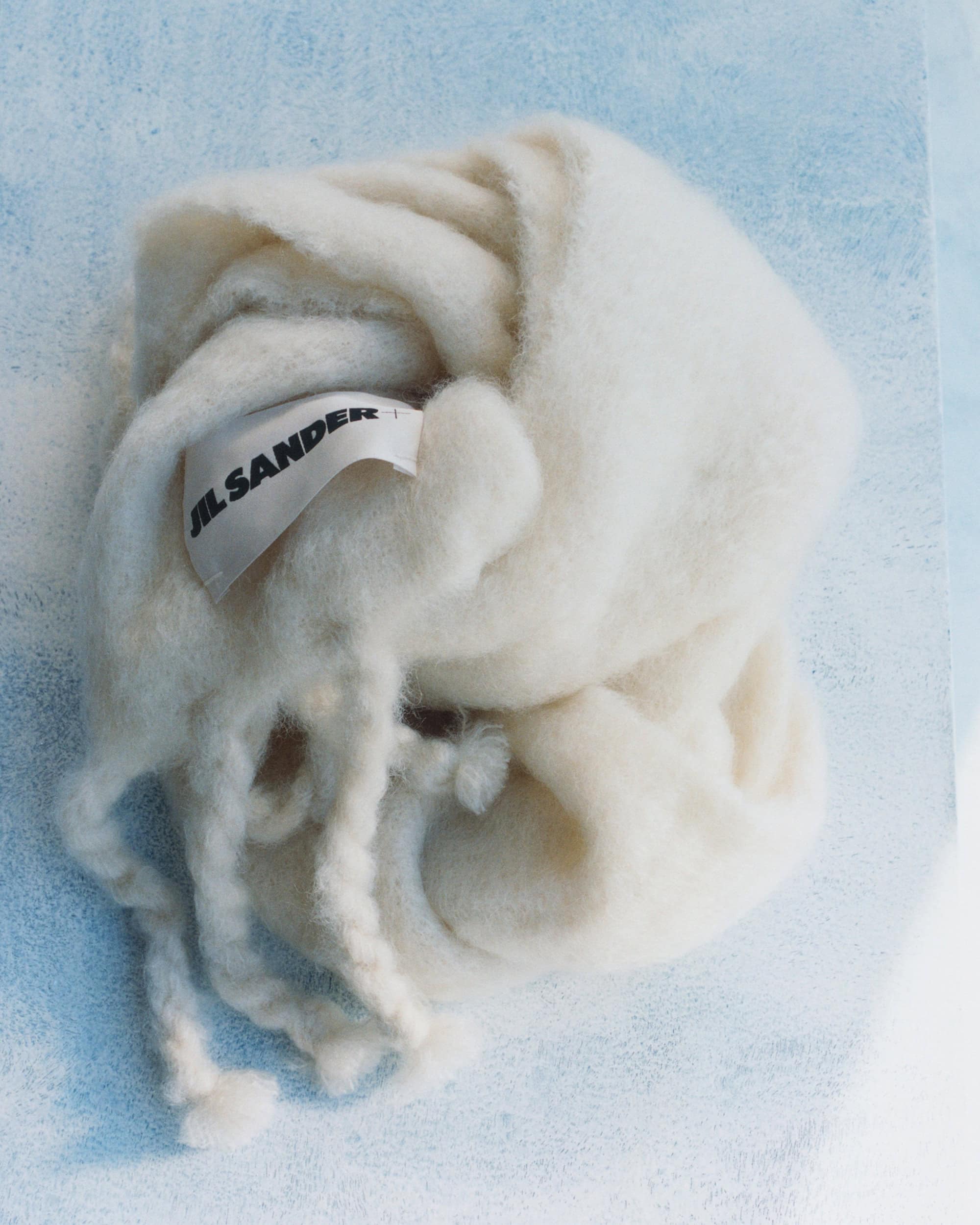 Soft cream-colored woolen scarf with visible branded label resting on a textured light blue and white surface.
