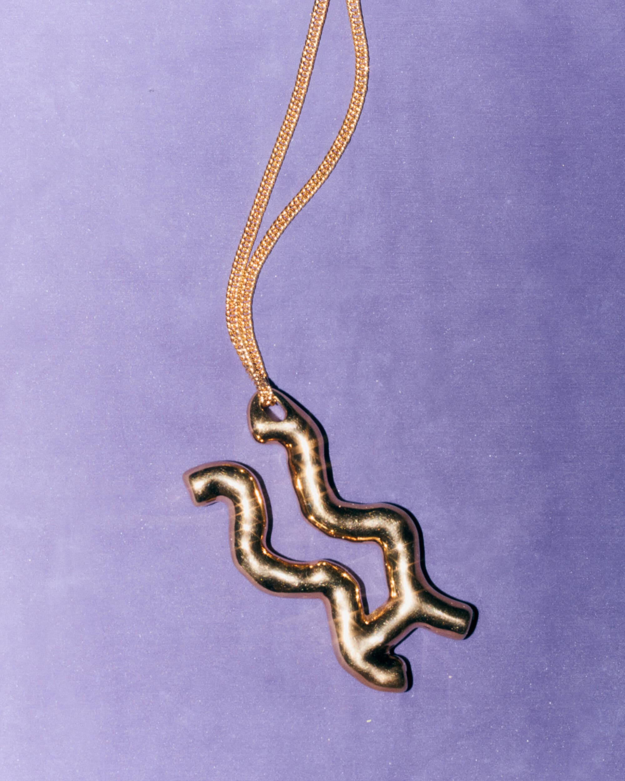 Close up of a sculptural gold pendant necklace hanging against a solid purple background