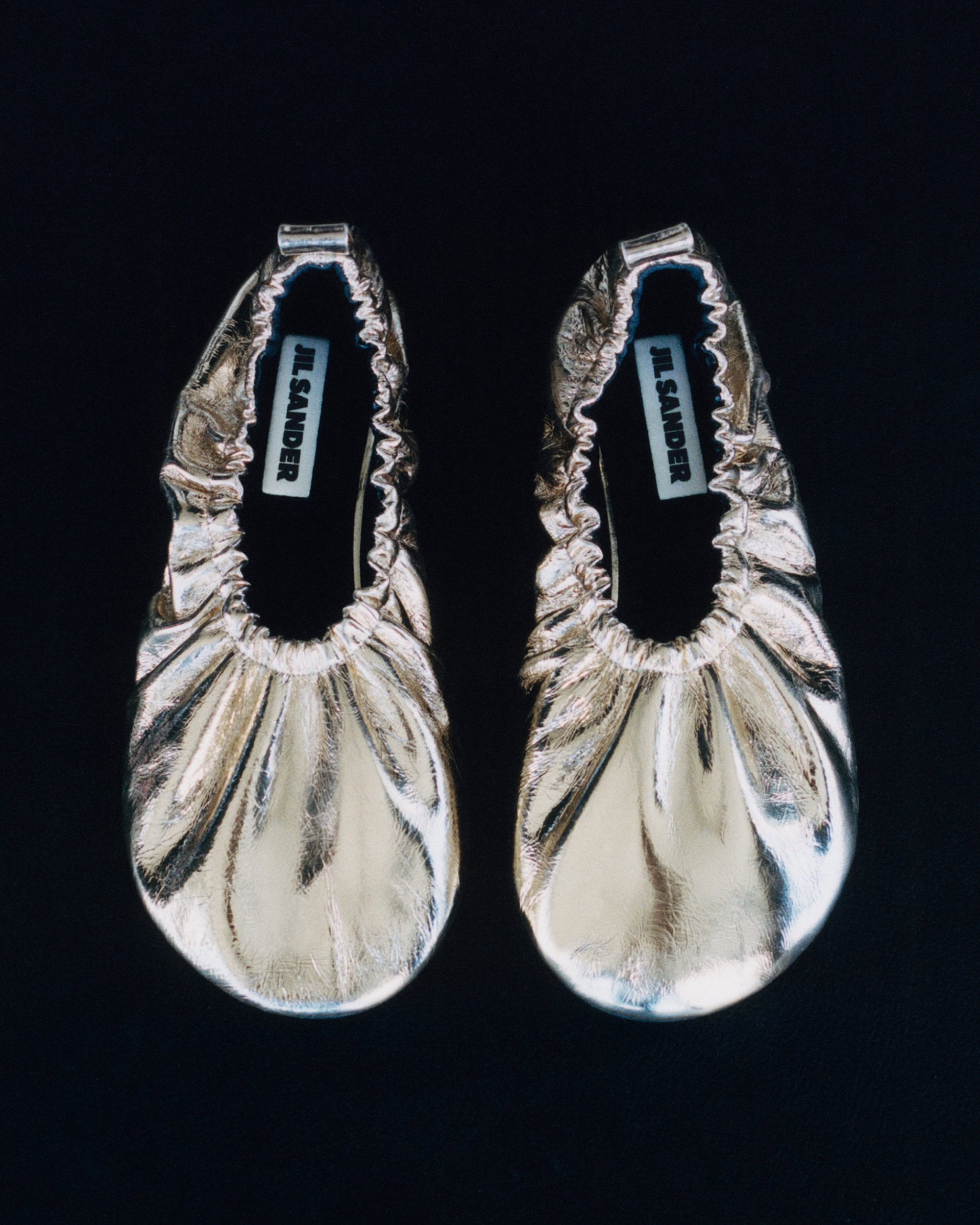 Pair of metallic gold leather flat shoes with gathered elastic rims on a solid black background.