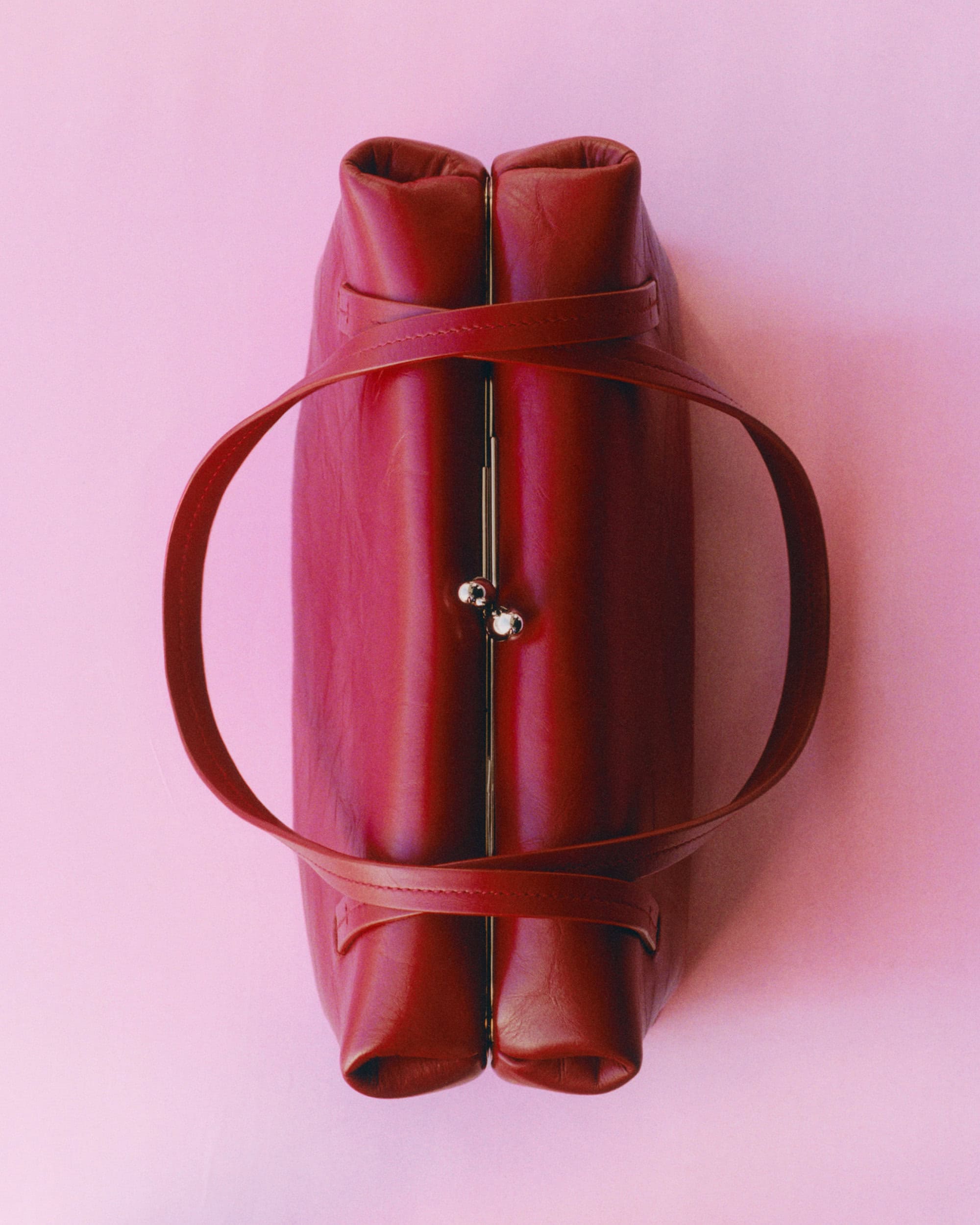 Top-down view of a structured red leather handbag with twin metal kiss-lock closures against a solid pink background.