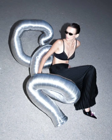 Model in black bra and pleated skirt seated next to a large metallic duct prop on asphalt.