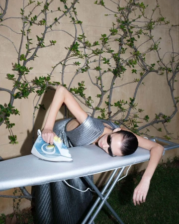 Model wearing dark sunglasses and a pleated silver dress lying across an ironing board outdoors