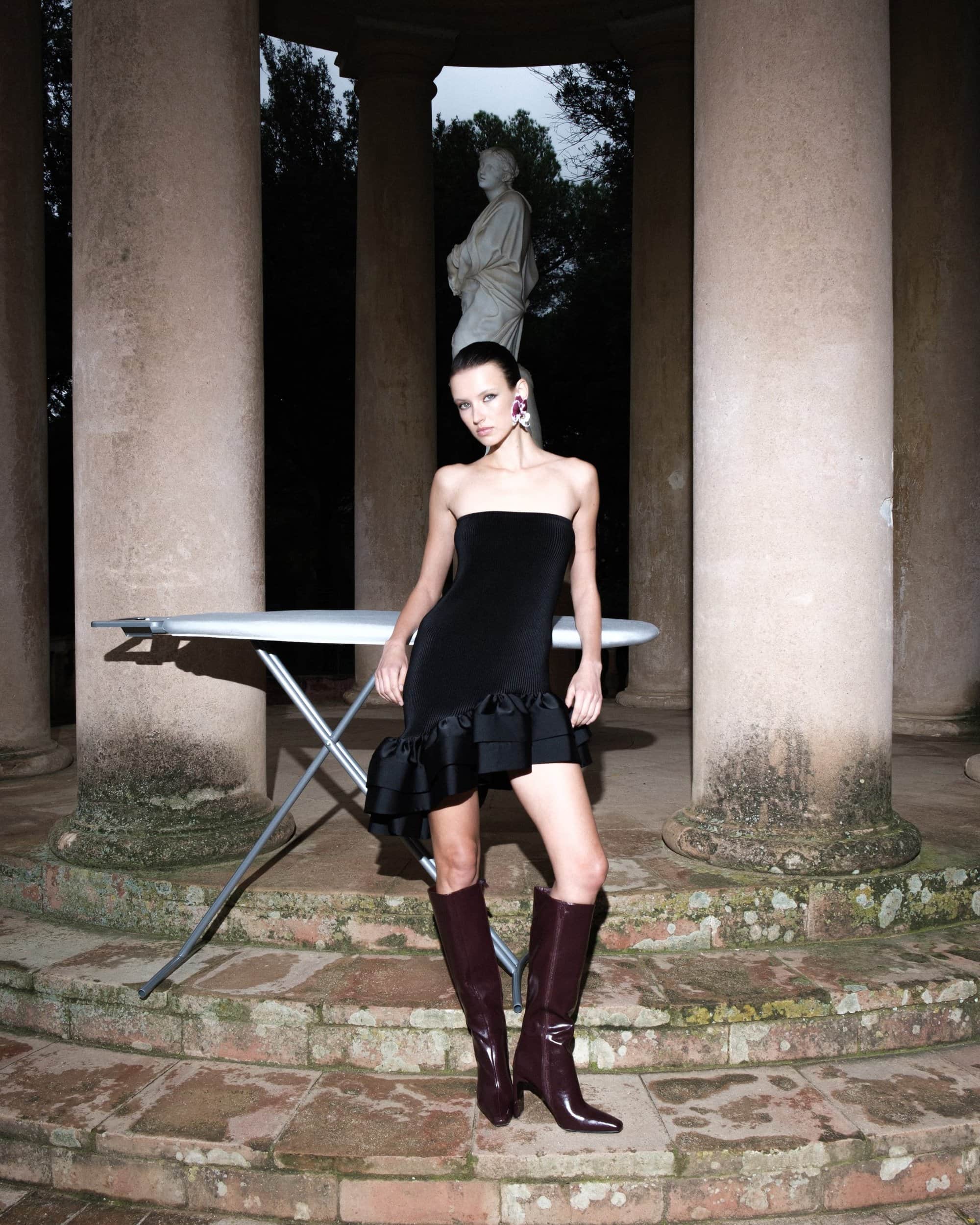 Model in strapless black dress and knee-high boots standing before classical columns and an ironing board for L'idee Woman Fall 2024 campaign.
