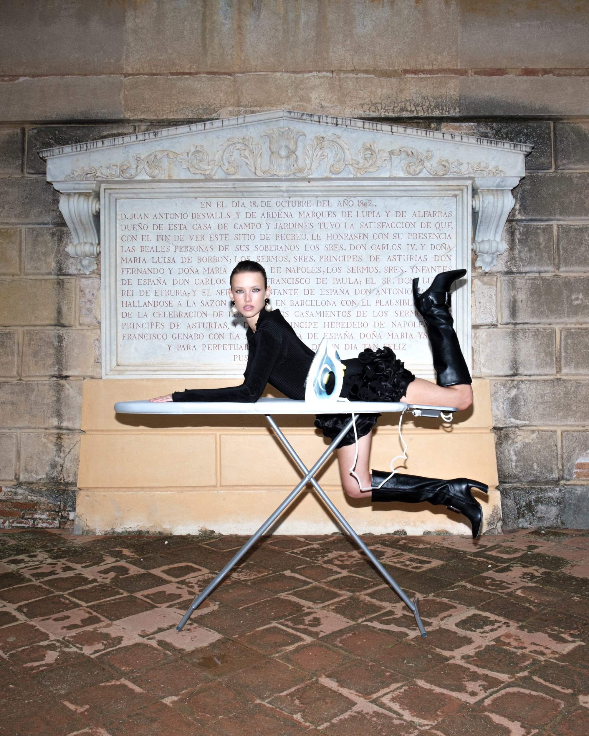 Model wearing black clothing and tall boots posed on an ironing board outdoors against an inscribed stone wall.