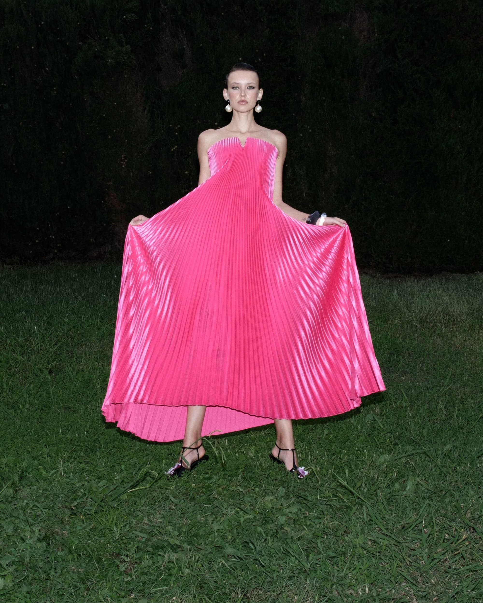 Model wearing a vibrant pink pleated strapless dress standing outdoors on a lawn at night for L'idee Woman Fall 2024 ad campaign.