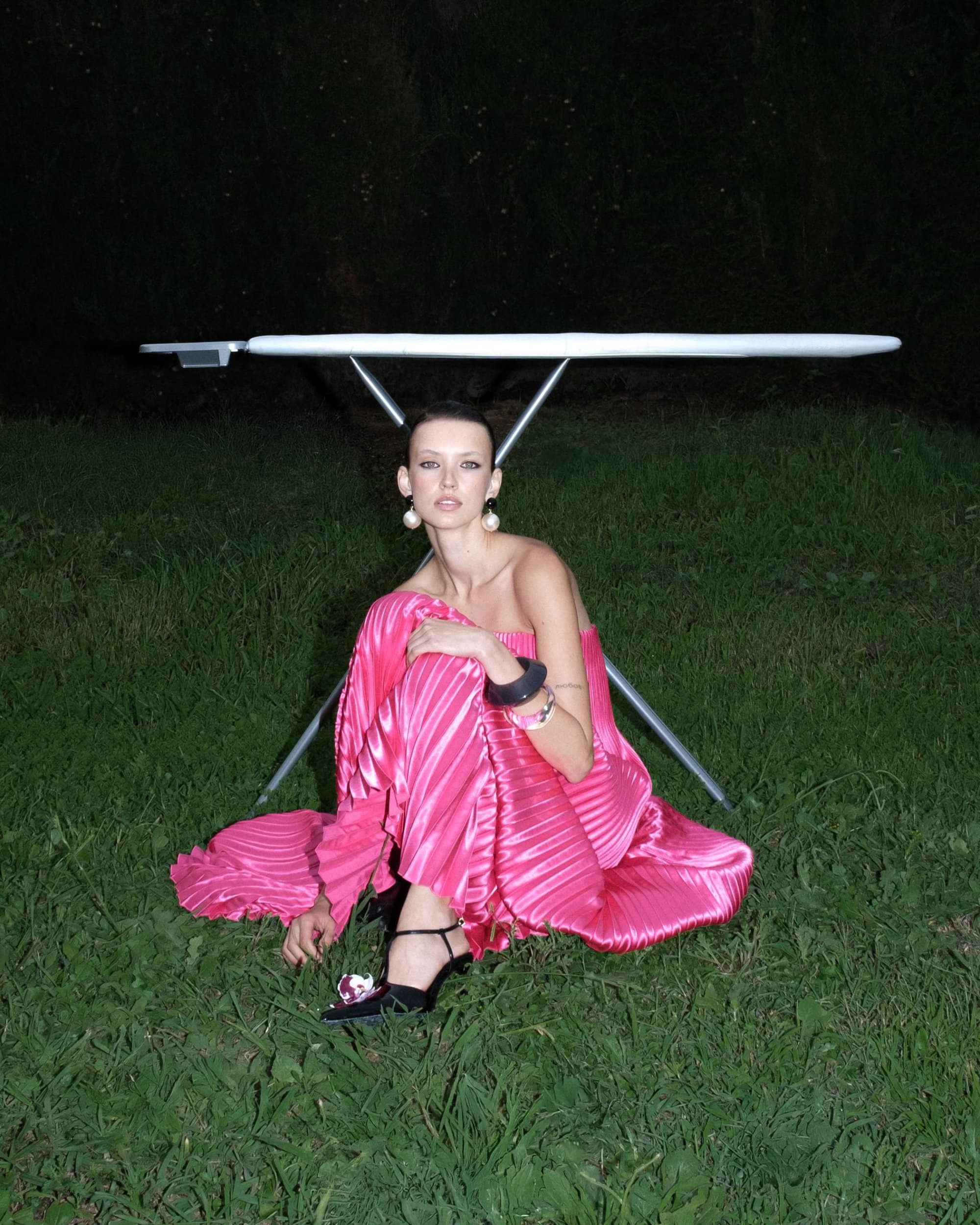 Model kneeling in dark grass wearing a pleated pink gown beneath a metal ironing board