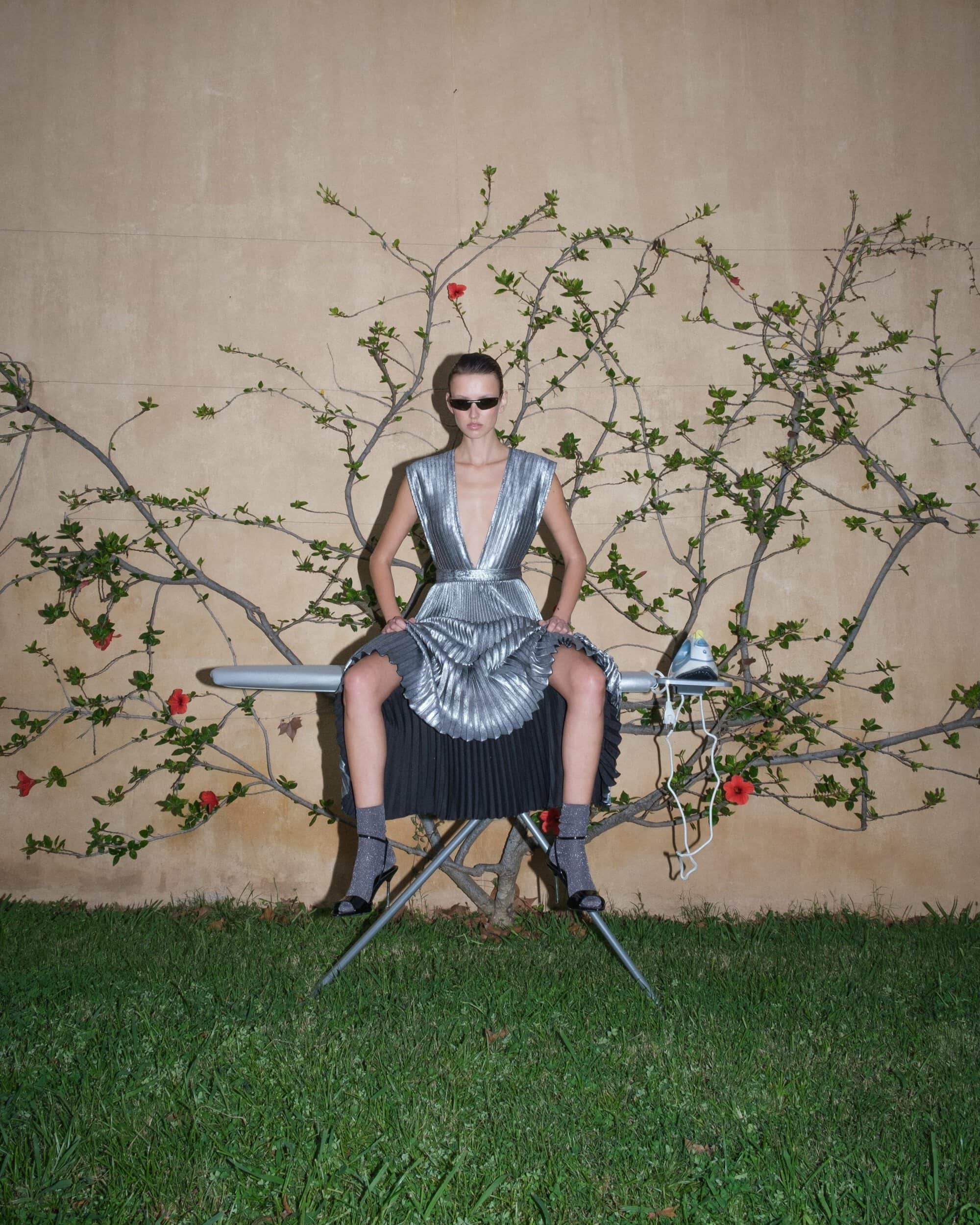 Model wearing a pleated silver metallic dress and sunglasses sitting on an ironing board outdoors