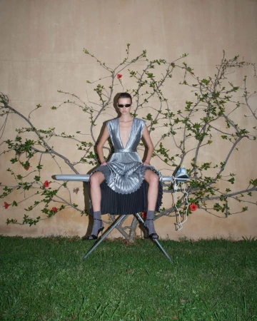 Model wearing a pleated silver metallic dress and sunglasses sitting on an ironing board outdoors