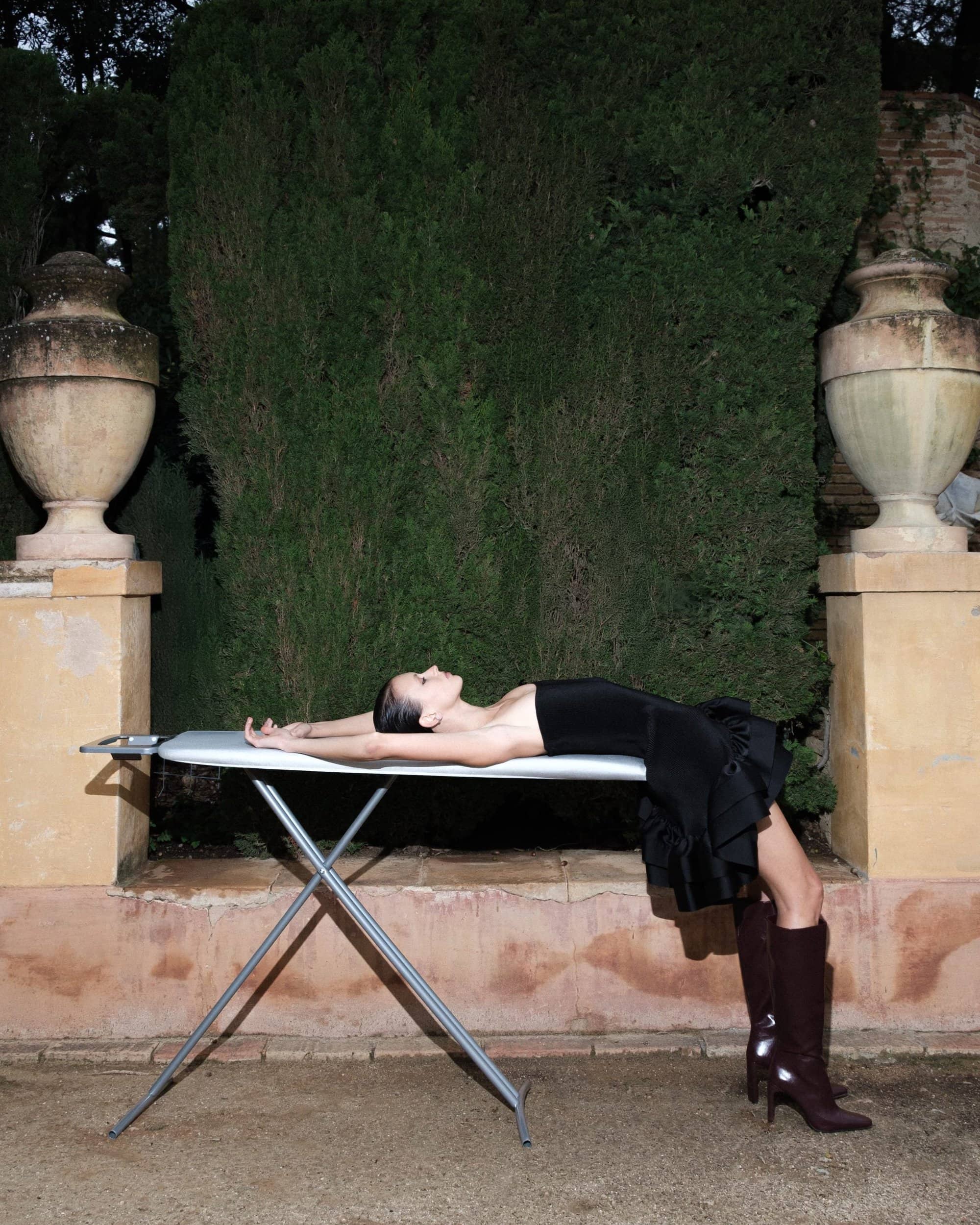 Model wearing a black strapless dress and dark boots lying back on an ironing board outdoors between stone urns