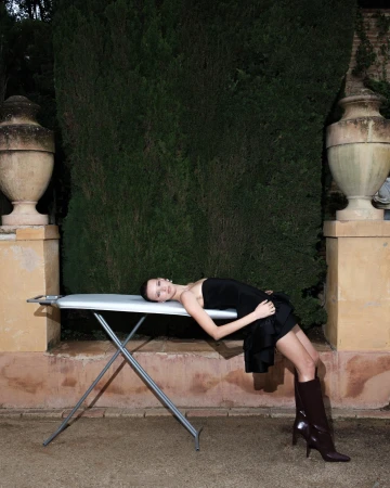 Model wearing black dress and tall brown boots lying across an ironing board outdoors