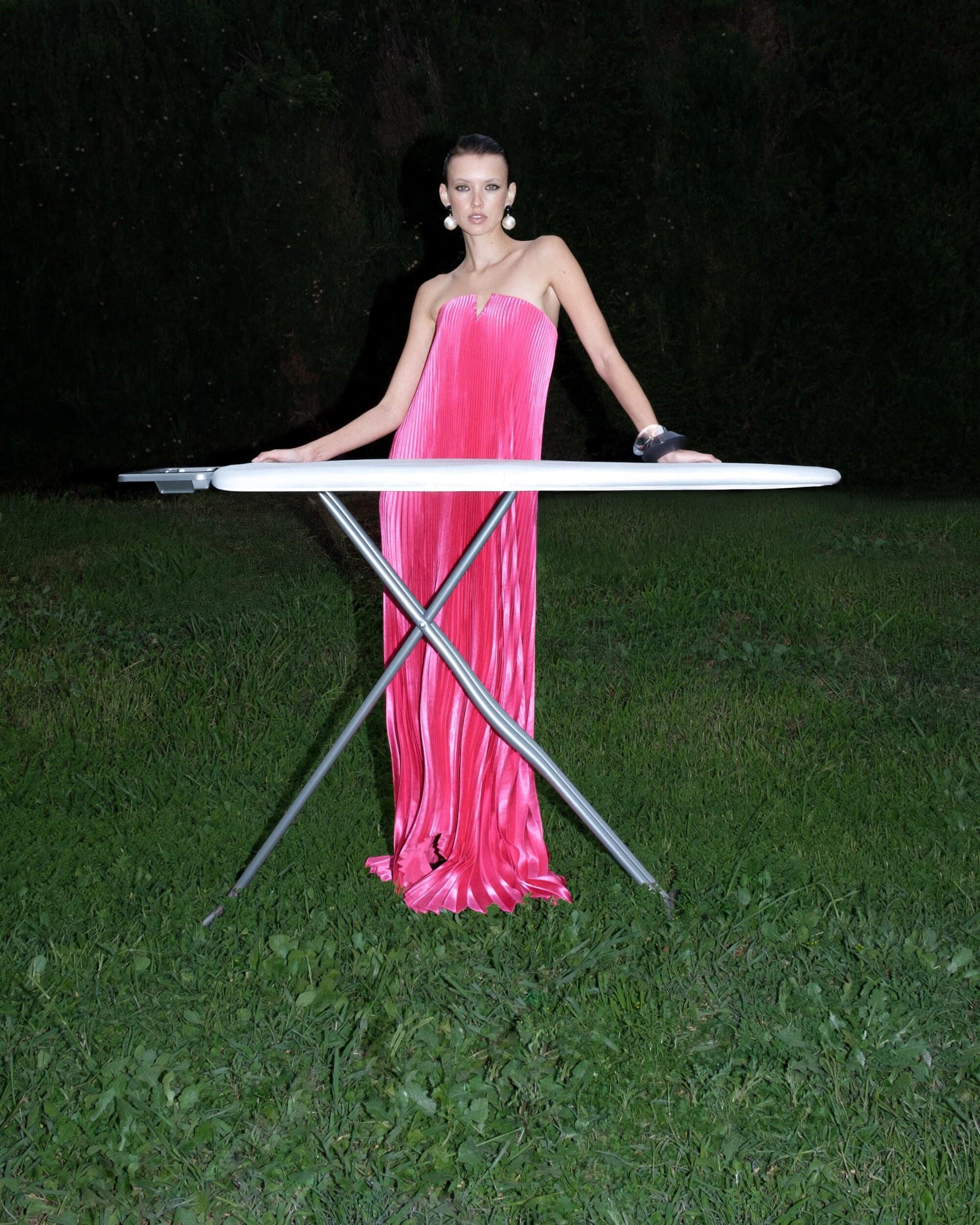 Model wearing a vibrant pink pleated strapless gown standing outdoors behind an ironing board at night.