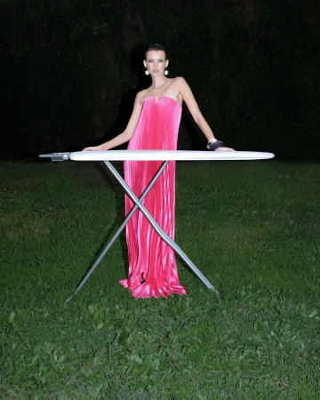 Model wearing a vibrant pink pleated strapless gown standing outdoors behind an ironing board at night.