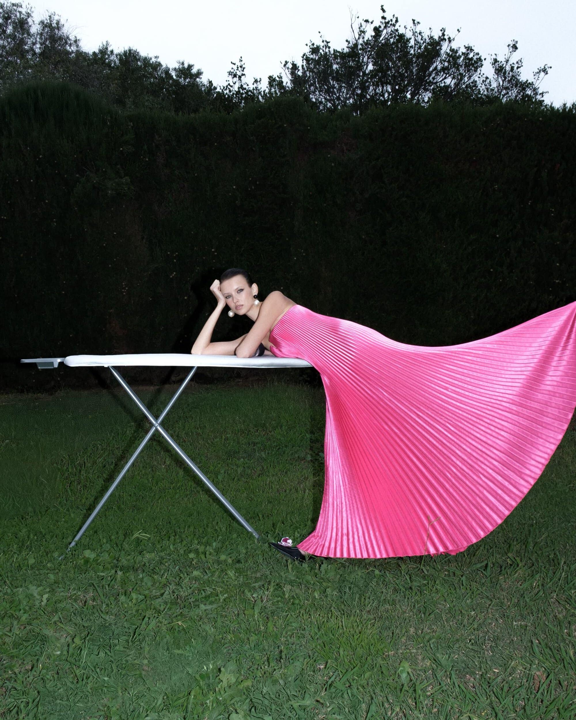 Model in a bright pink pleated gown leaning against an ironing board outdoors on a lawn at night