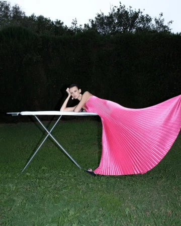 Model in a bright pink pleated gown leaning against an ironing board outdoors on a lawn at night